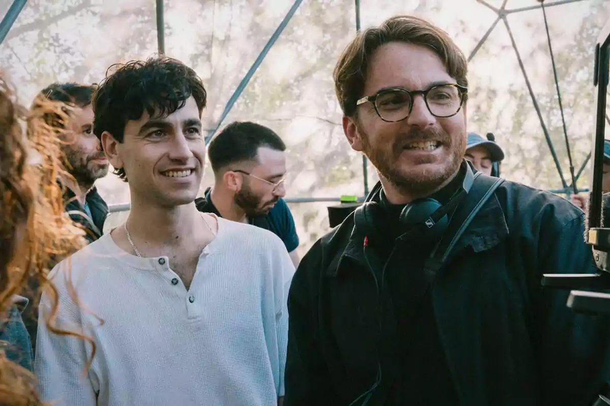 Alex Monner junto al director de La Ruta sonríe mientras observa algo fuera de cámara en un entorno al aire libre.