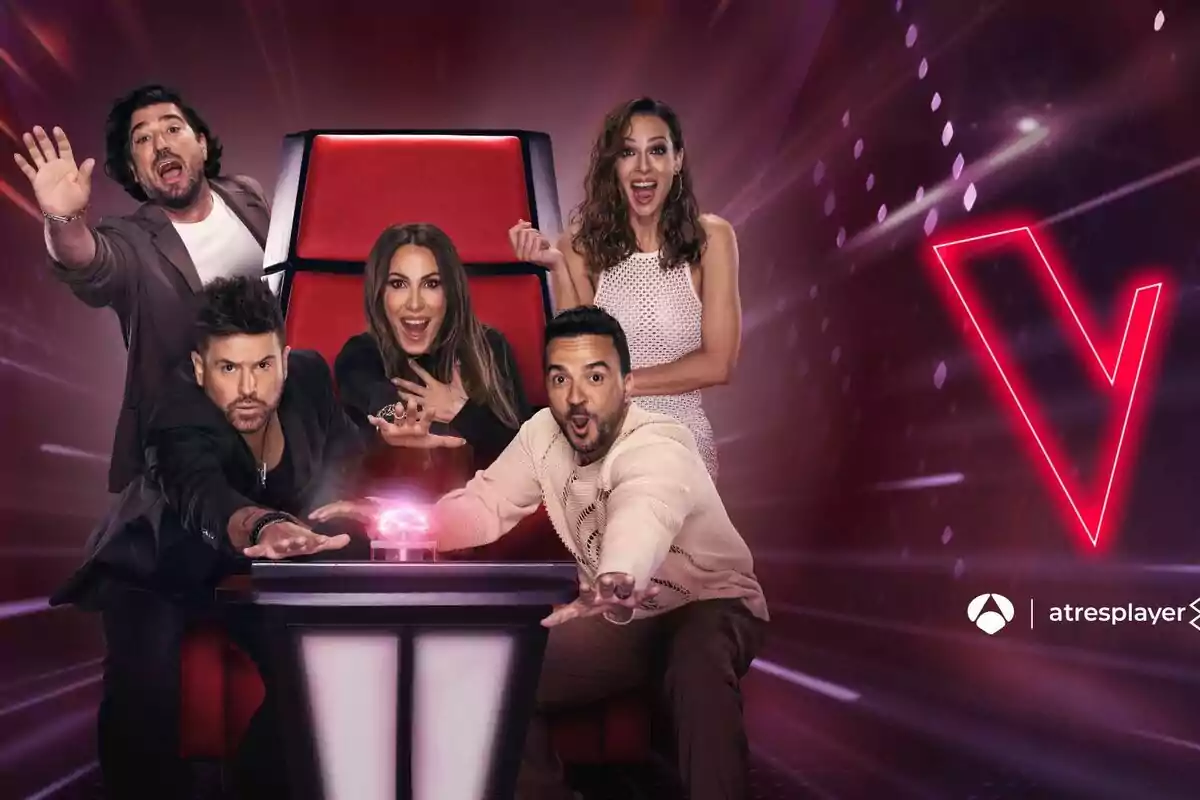 Eva González, Antonio Orozco, Malú, Luis Fonsi y Pablo López posan emocionadas alrededor de una silla roja con un botón iluminado en el centro, con un fondo oscuro y un logotipo de La Voz en la esquina superior derecha.