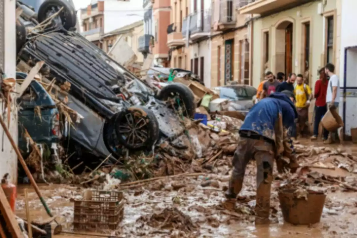 Una calle llena de escombros y barro tras una inundación con un coche volcado y personas limpiando.