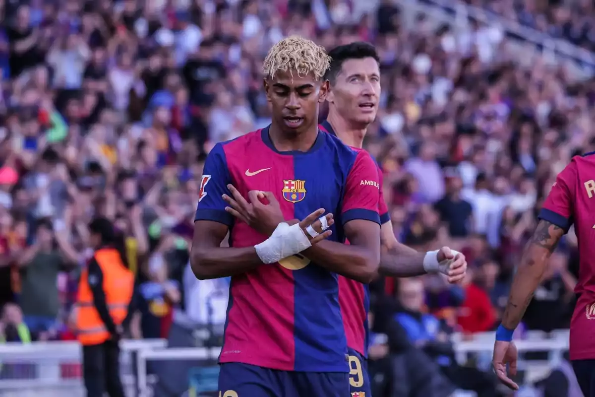 Un jugador de fútbol con el uniforme del FC Barcelona celebra en el campo mientras otro jugador está detrás de él y el público observa desde las gradas. Un jugador de fútbol con el uniforme del FC Barcelona celebra en el campo mientras otro jugador está detrás de él y el público observa desde las gradas.