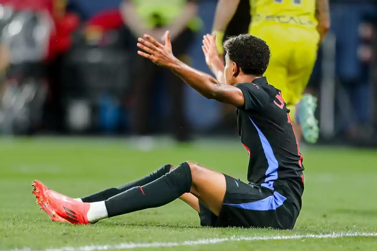 Jugador de fútbol sentado en el césped con los brazos levantados durante un partido.