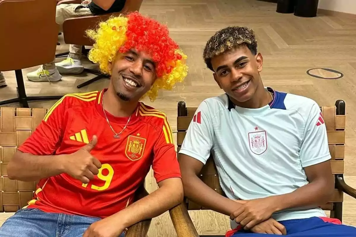 Lamine Yamal y su padre, sentados en sillas, el padre con una peluca de colores rojo y amarillo y una camiseta roja de la selección española de fútbol, y el hijo con una camiseta blanca de la misma selección.