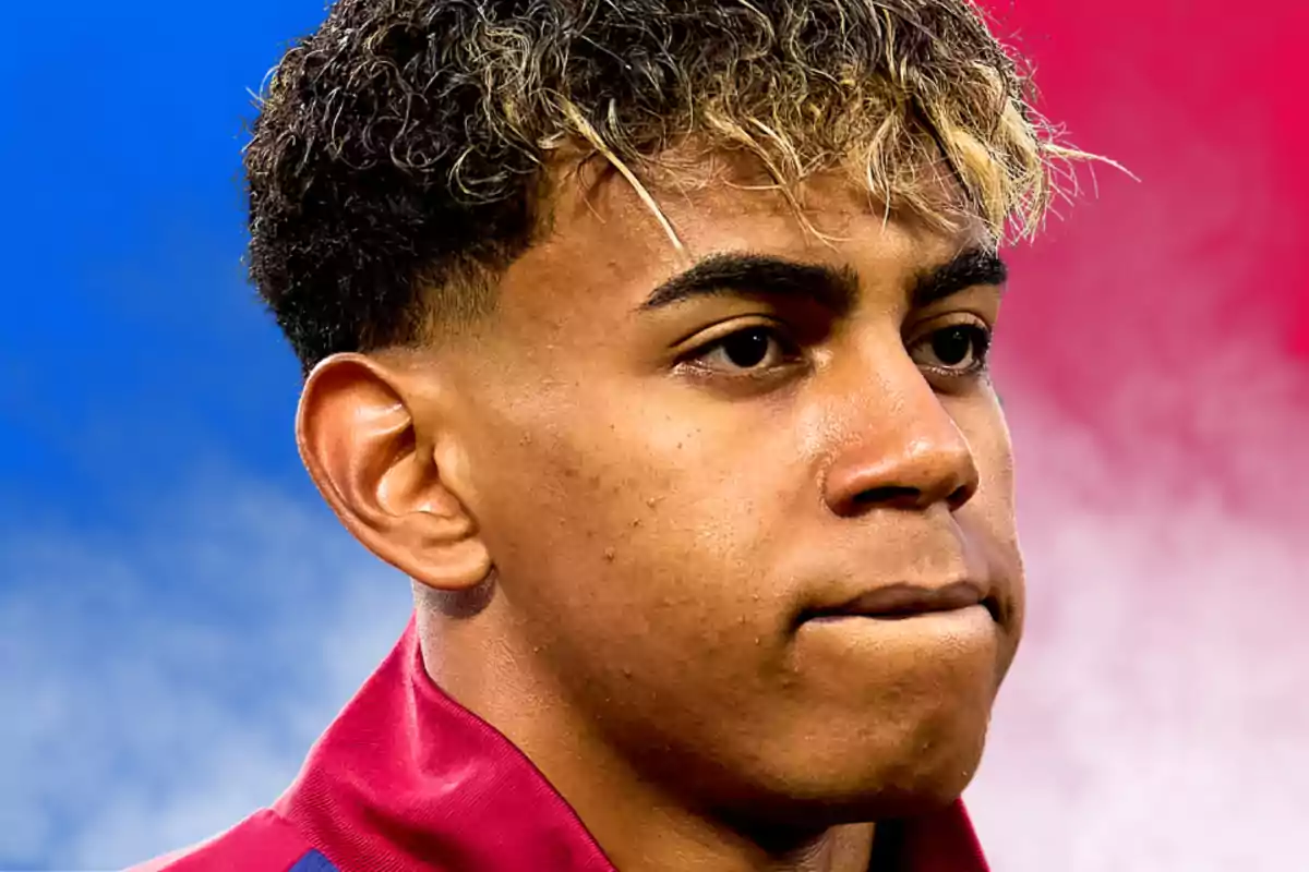 Joven con cabello rizado y puntas rubias mirando hacia el frente con expresión seria, fondo difuminado en tonos azul y rosa.