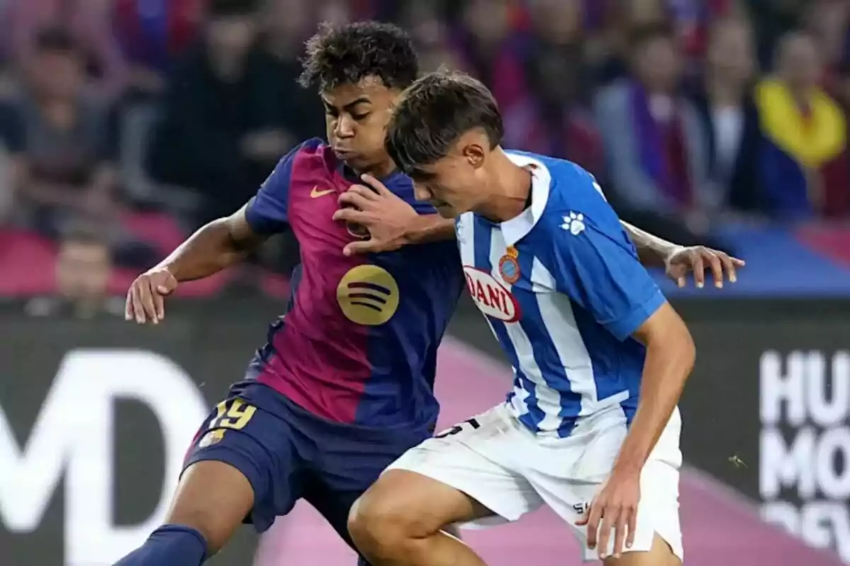Lamine Yamal y otro jugador de fútbol compiten por el balón en un partido, uno viste un uniforme azul y granate del Barça y el otro un uniforme azul y blanco del Espanyol.