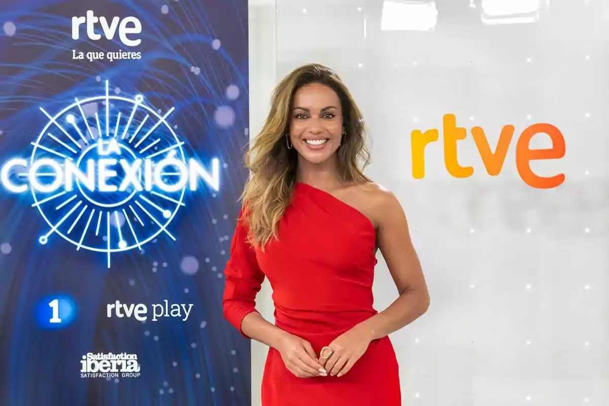 Lara Álvarez con vestido rojo de un solo hombro sonriendo frente a un fondo con los logotipos de RTVE y La Conexión