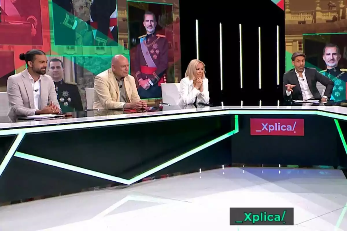 Un grupo de personas encabezadas por José Yélamo sentadas en un set de televisión con pantallas de fondo mostrando imágenes de una figura en uniforme militar y el logo del programa _Xplíca! en el escritorio.