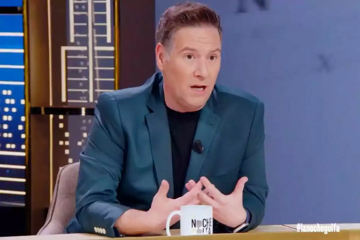 Carlos Latre con chaqueta azul oscuro está sentado en un set de televisión, gesticulando mientras habla y tiene una taza blanca sobre la mesa en 'La noche golfa'.