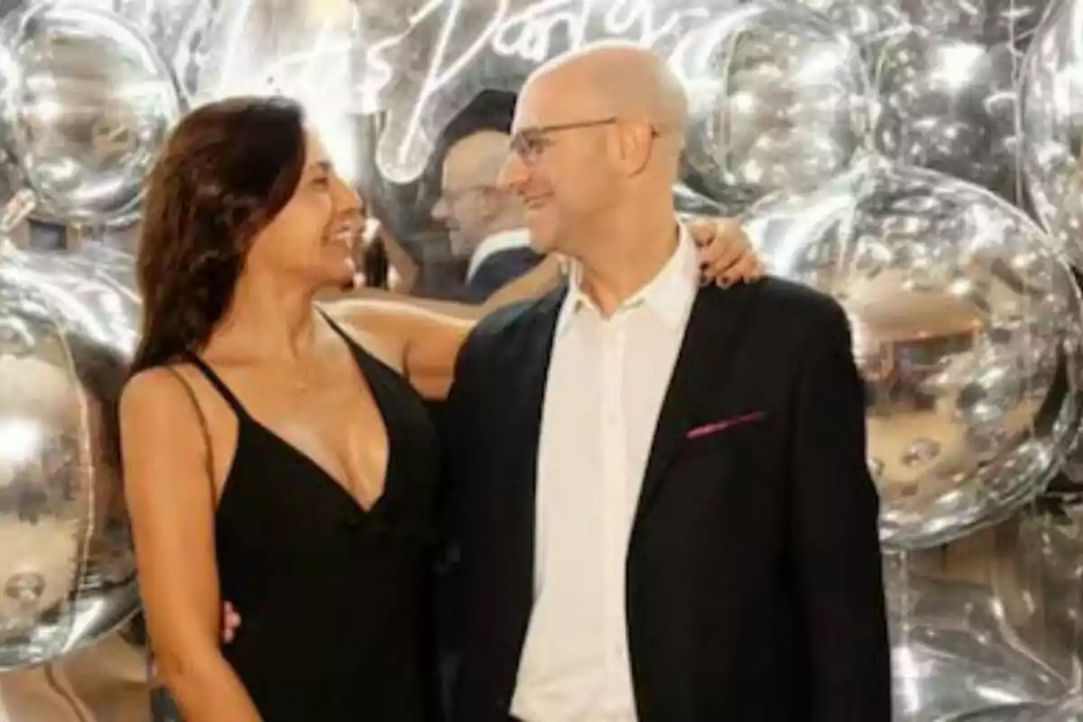 Una pareja sonriente se mira con afecto mientras posa frente a un fondo de globos plateados. Una pareja sonriente se mira con afecto mientras posa frente a un fondo de globos plateados.
