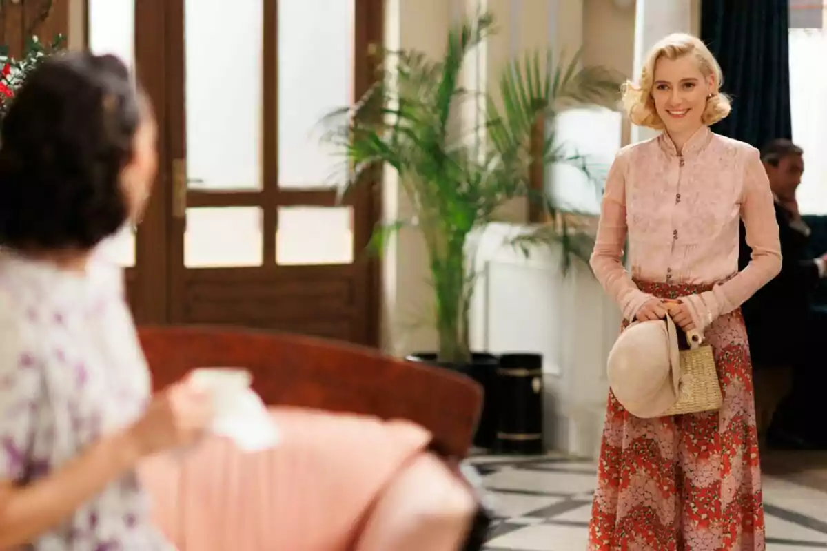 Laurita rubia con una blusa rosa y una falda larga estampada sostiene un sombrero y una bolsa de mimbre mientras sonríe en un interior elegante con plantas y muebles clásicos en La Moderna.