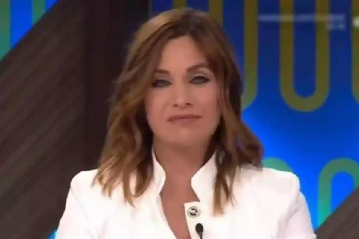 Leire Martínez con cabello castaño y ondulado lleva una chaqueta blanca y está frente a un fondo azul con detalles amarillos.