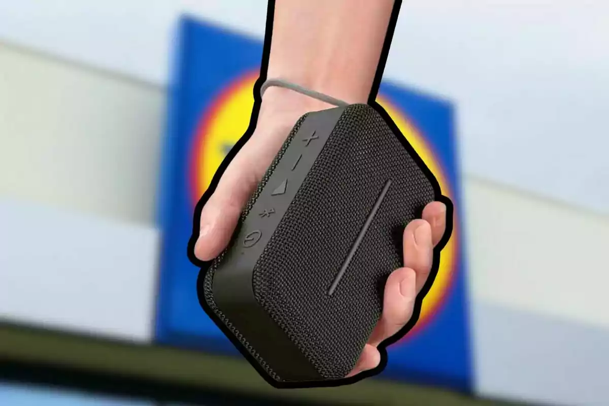 Una mano sosteniendo un altavoz portátil negro con botones de control visibles y un fondo borroso de un edificio con un letrero azul y amarillo. Una mano sosteniendo un altavoz portátil negro con botones de control visibles y un fondo borroso de un edificio con un letrero azul y amarillo.