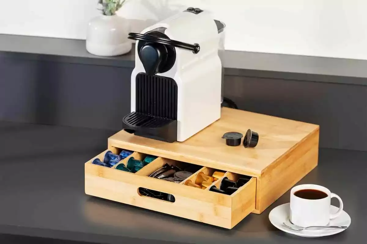 Cafetera de cápsulas blanca sobre una caja organizadora de madera con cápsulas de café y una taza de café en una mesa Cafetera de cápsulas blanca sobre una caja organizadora de madera con cápsulas de café y una taza de café en una mesa