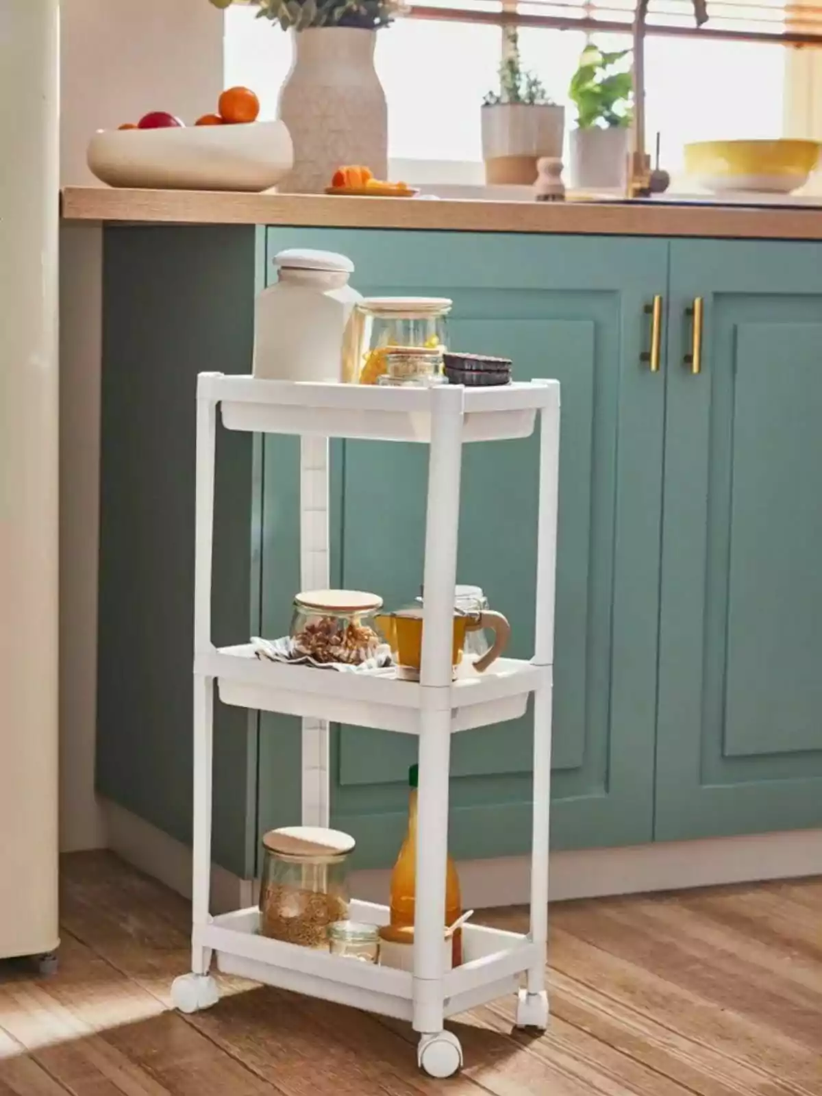 Un carrito de cocina blanco con ruedas sostiene varios frascos y botellas, ubicado frente a un mueble verde con encimera de madera en una cocina iluminada. Un carrito de cocina blanco con ruedas sostiene varios frascos y botellas, ubicado frente a un mueble verde con encimera de madera en una cocina iluminada.