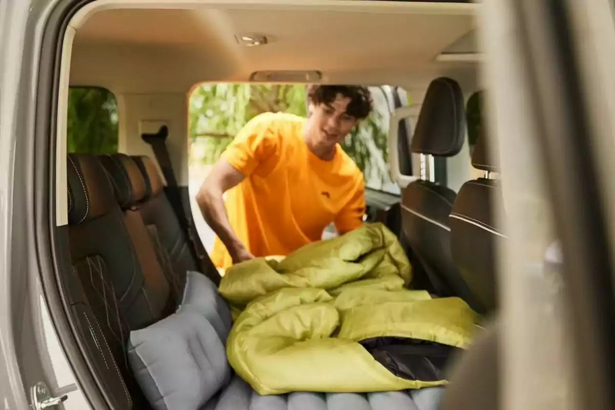 Un hombre con camiseta naranja acomoda un saco de dormir verde en el interior de un vehículo. Un hombre con camiseta naranja acomoda un saco de dormir verde en el interior de un vehículo.