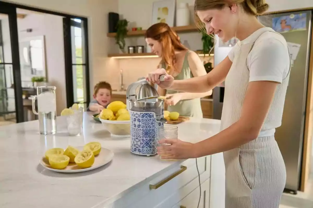 Una persona exprime limones en una cocina moderna mientras otra persona y un niño están en el fondo, con un plato de limones cortados y una jarra de agua en la encimera. Una persona exprime limones en una cocina moderna mientras otra persona y un niño están en el fondo, con un plato de limones cortados y una jarra de agua en la encimera.