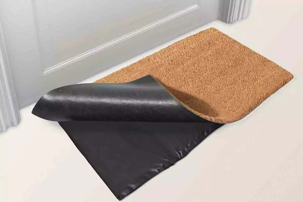 Un felpudo marrón con la esquina levantada mostrando una capa negra antideslizante debajo, colocado frente a una puerta blanca. Un felpudo marrón con la esquina levantada mostrando una capa negra antideslizante debajo, colocado frente a una puerta blanca.