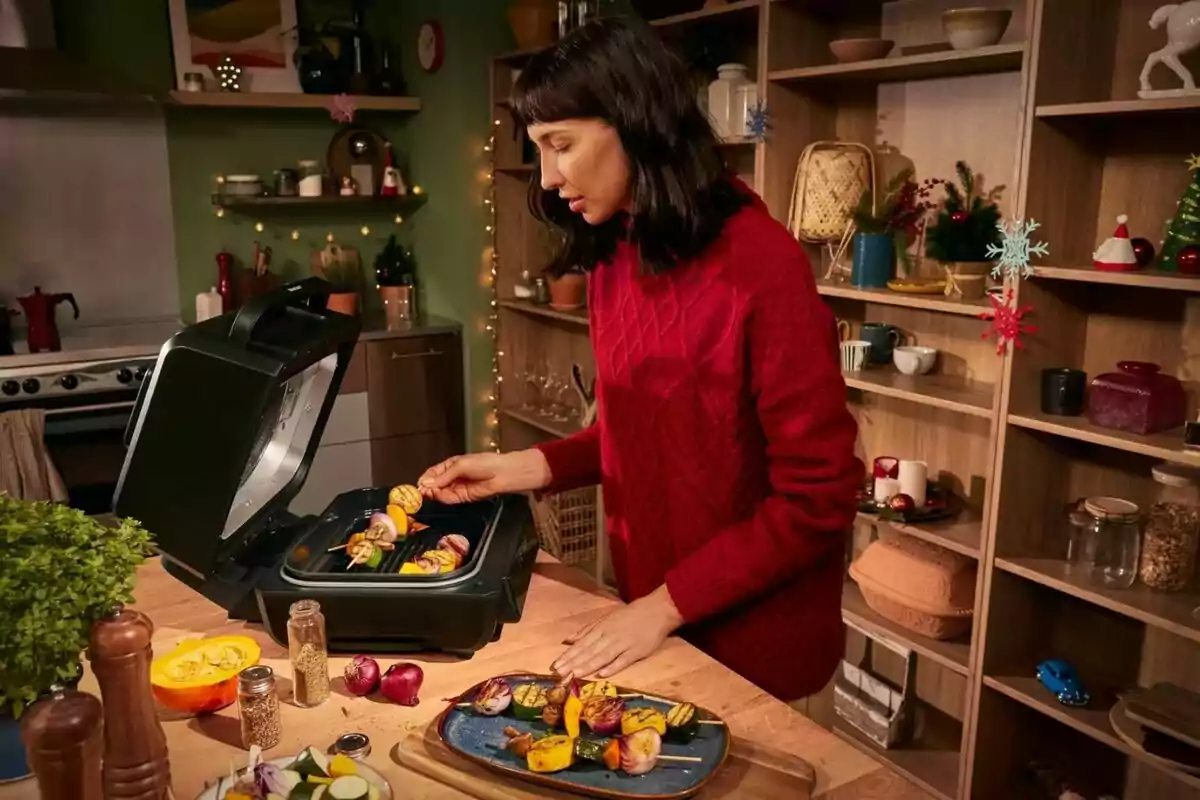 Una persona cocinando brochetas de verduras en una parrilla eléctrica en una cocina decorada con luces y adornos festivos. Una persona cocinando brochetas de verduras en una parrilla eléctrica en una cocina decorada con luces y adornos festivos.
