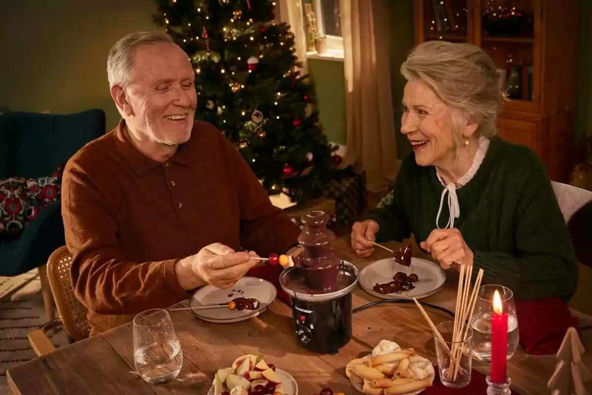 Una pareja de personas mayores sonríe mientras disfruta de una fuente de chocolate en una mesa decorada para Navidad con un árbol iluminado al fondo. Una pareja de personas mayores sonríe mientras disfruta de una fuente de chocolate en una mesa decorada para Navidad con un árbol iluminado al fondo.