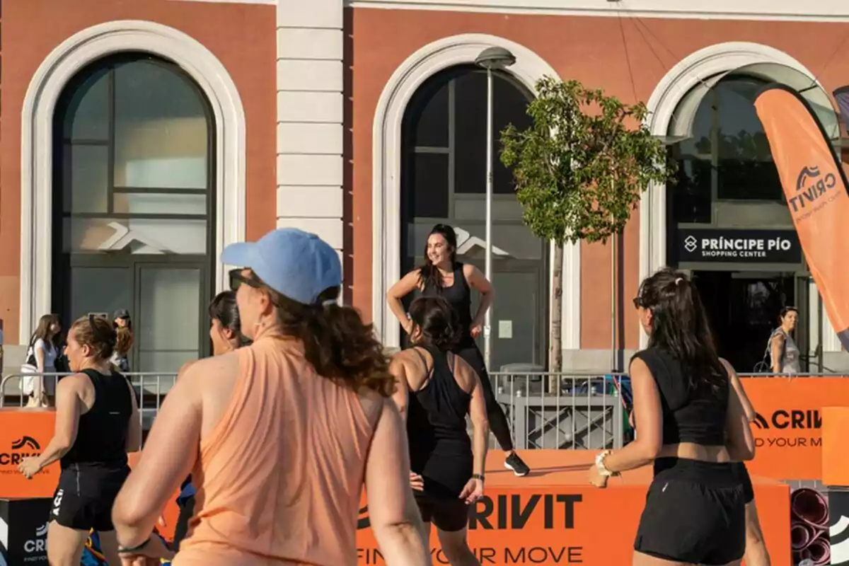 Un grupo de mujeres hace ejercicio al aire libre frente a un edificio con grandes ventanas y un letrero de centro comercial mientras una instructora sonríe sobre una tarima Un grupo de mujeres hace ejercicio al aire libre frente a un edificio con grandes ventanas y un letrero de centro comercial mientras una instructora sonríe sobre una tarima