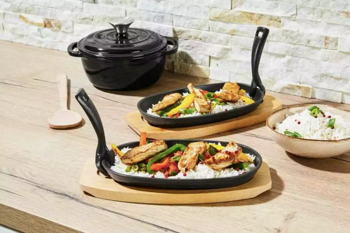 Dos sartenes con arroz, pollo y vegetales sobre una mesa de madera, junto a una olla negra con tapa y una cuchara de madera. Dos sartenes con arroz, pollo y vegetales sobre una mesa de madera, junto a una olla negra con tapa y una cuchara de madera.