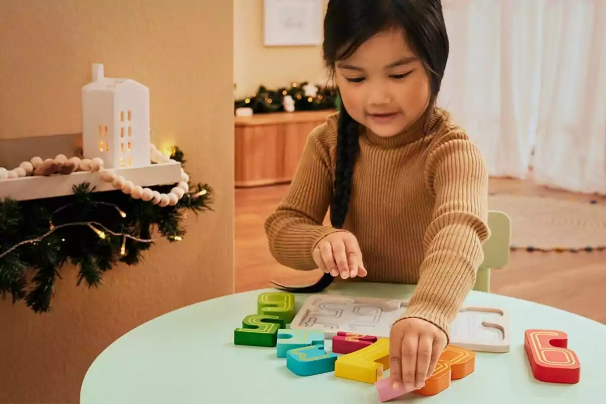 Una niña juega con un rompecabezas de números de colores en una mesa mientras una decoración navideña adorna la pared cercana. Una niña juega con un rompecabezas de números de colores en una mesa mientras una decoración navideña adorna la pared cercana.
