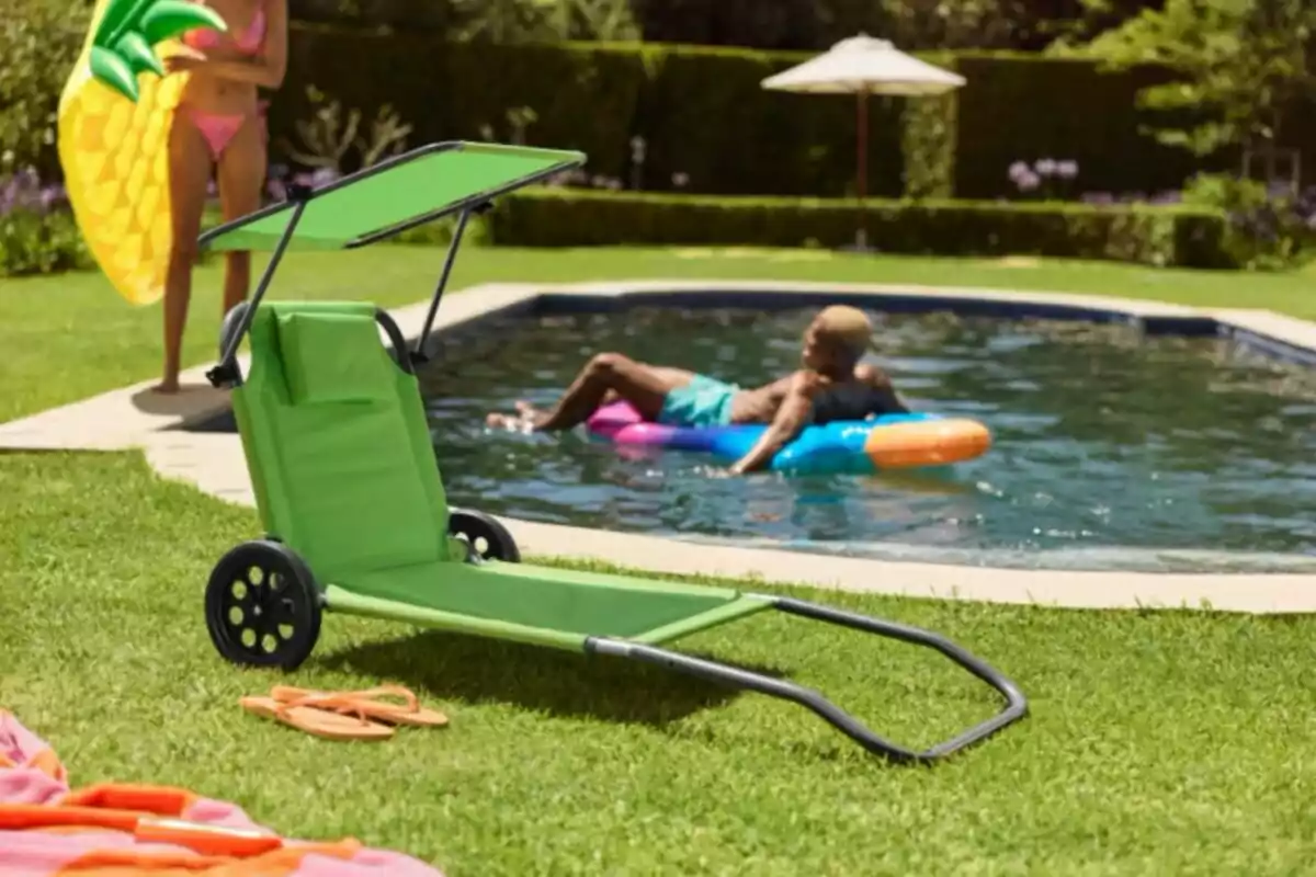 Silla verde con ruedas y toldo en el césped junto a una piscina donde una persona flota en un inflable y otra sostiene un colchón de piña Silla verde con ruedas y toldo en el césped junto a una piscina donde una persona flota en un inflable y otra sostiene un colchón de piña