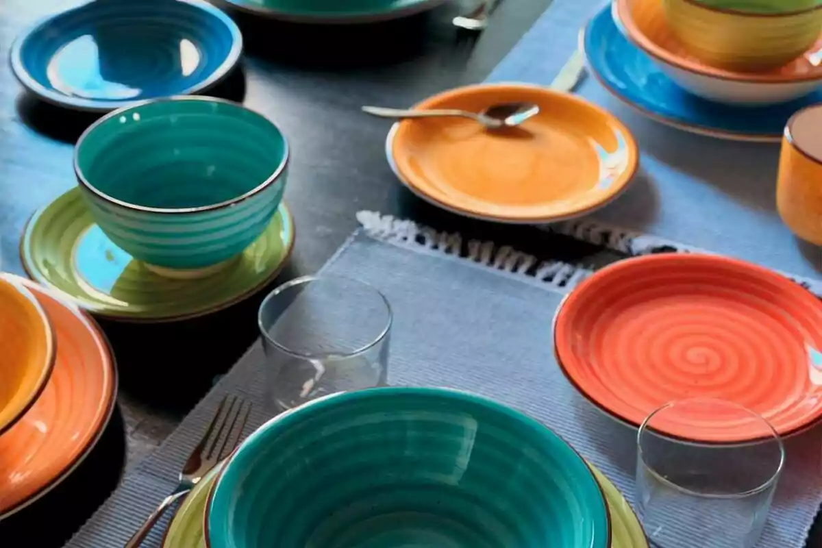 Vajilla colorida con platos y tazones en tonos de azul, naranja y verde sobre un mantel azul con cubiertos y vasos. Vajilla colorida con platos y tazones en tonos de azul, naranja y verde sobre un mantel azul con cubiertos y vasos.