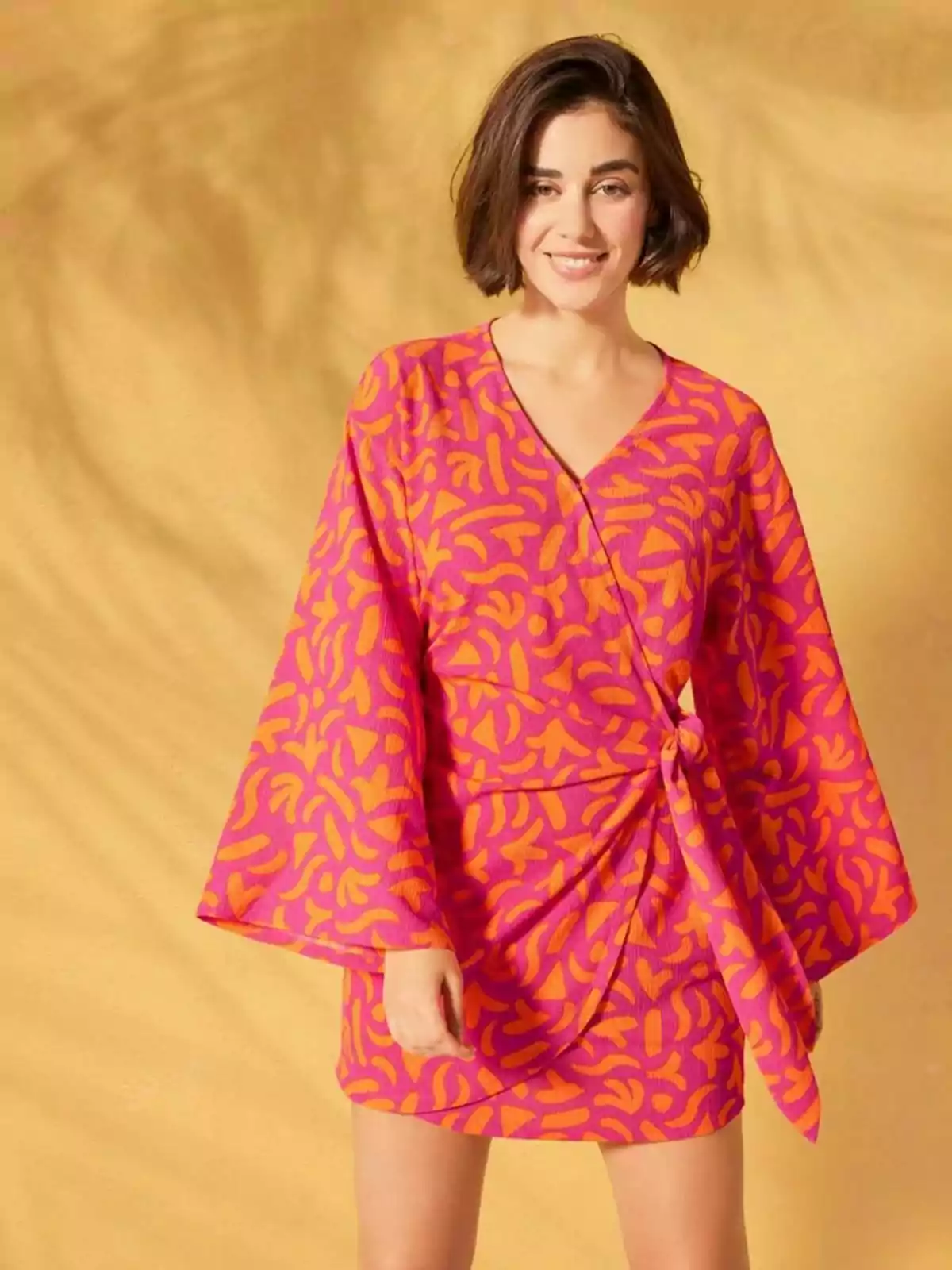 Una persona con cabello corto y oscuro lleva un vestido colorido con estampado abstracto en tonos rosas y naranjas, posando frente a un fondo amarillo. Una persona con cabello corto y oscuro lleva un vestido colorido con estampado abstracto en tonos rosas y naranjas, posando frente a un fondo amarillo.