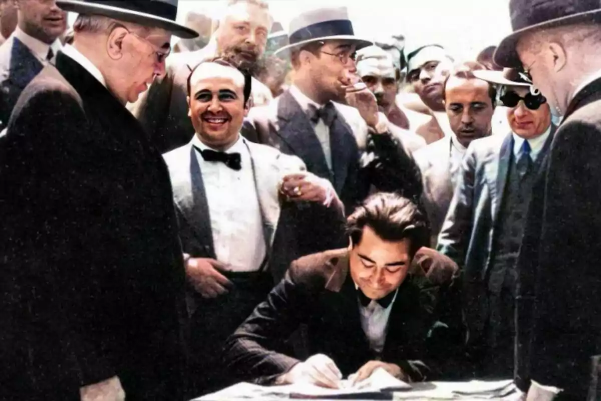Un grupo de hombres vestidos con trajes formales rodea a un hombre que está sentado y firmando un documento. Un grupo de hombres vestidos con trajes formales rodea a un hombre que está sentado y firmando un documento.