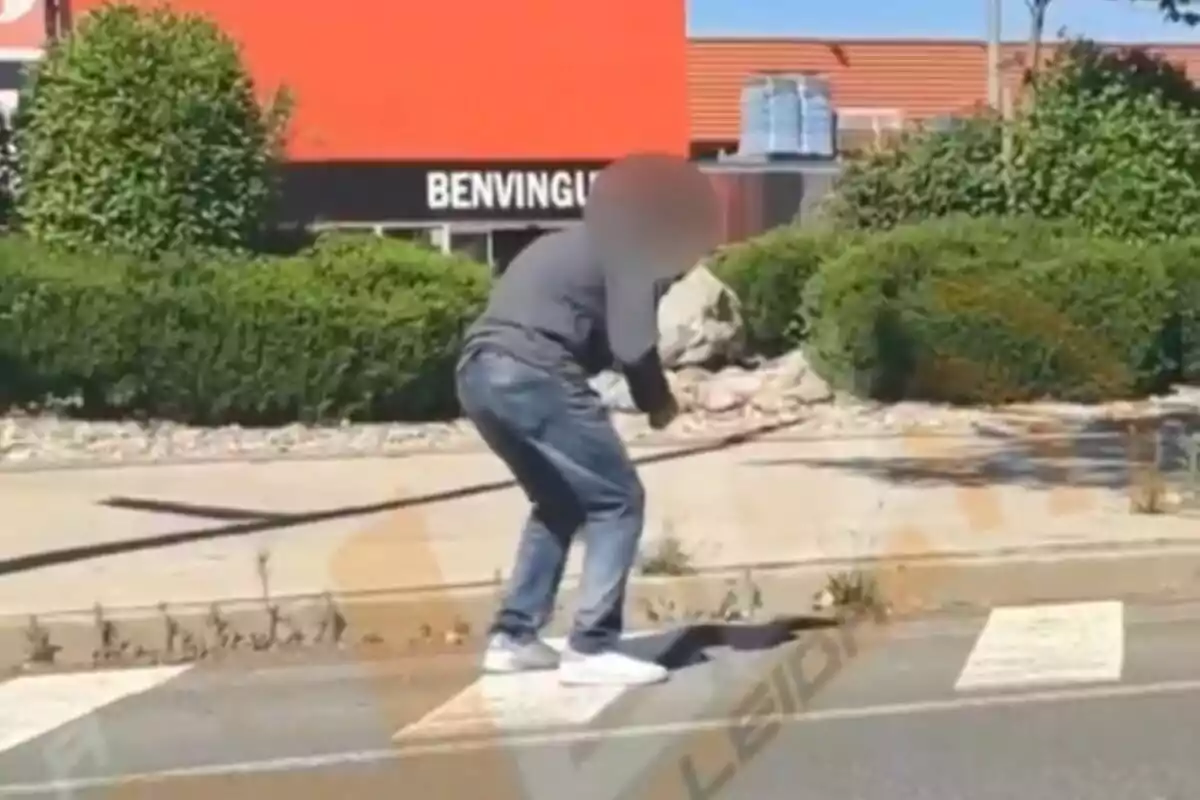 Una persona con el rostro difuminado está agachada en una acera cerca de un paso de peatones, con un fondo de arbustos y un edificio naranja que tiene un cartel que dice "BENVINGUTS".