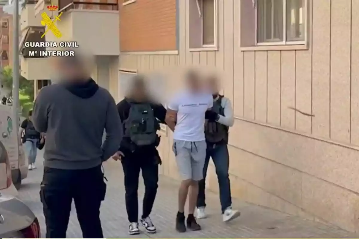 Varios agentes de la Guardia Civil escoltan a una persona esposada por una acera junto a un edificio, con los rostros de los presentes difuminados.