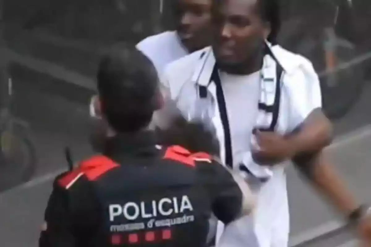 Un agente de policía con uniforme negro y rojo interactúa con dos personas en la calle