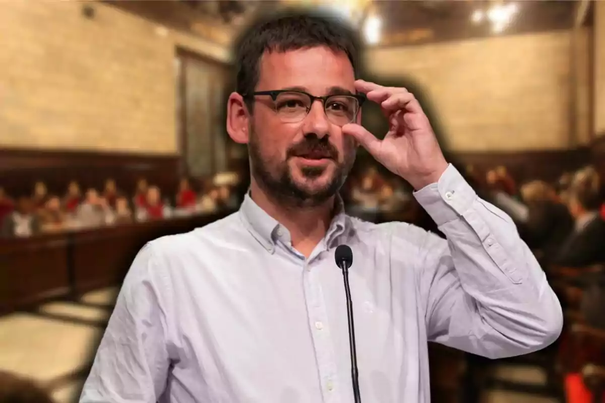 Un hombre con gafas y barba corta está de pie frente a un micrófono, ajustándose las gafas con una mano. Lleva una camisa blanca y parece estar en una sala de reuniones o conferencia, con varias personas desenfocadas en el fondo.