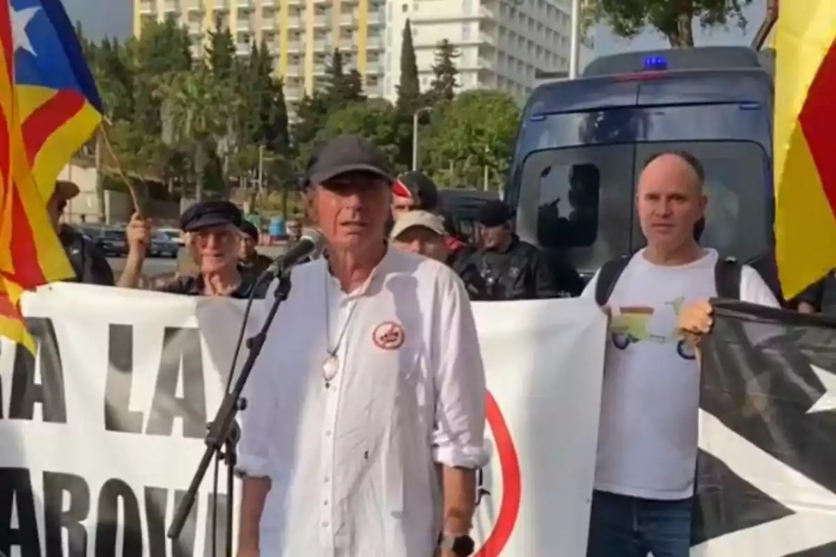 Un hombre con camisa blanca y gorra negra habla frente a un micrófono en una manifestación, rodeado de personas que sostienen banderas y pancartas.