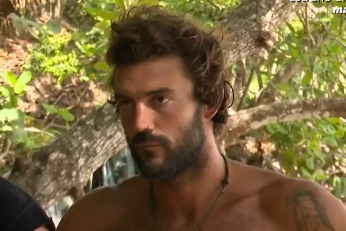 Captura de Logan Sampedro con barba y cabello rizado se encuentra al aire libre, rodeado de vegetación, en la isla de Supervivientes, con una expresión seria en su rostro.