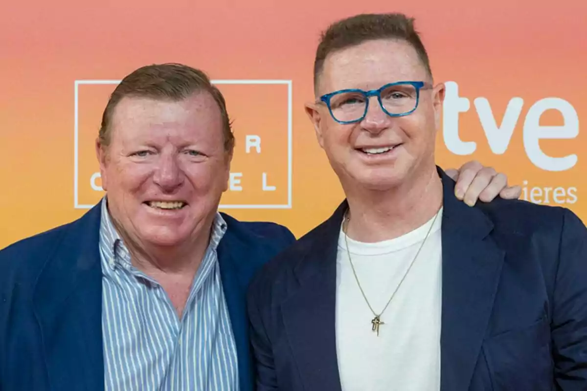 Jorge y César Cadaval, Los Morancos, sonrientes posando juntos frente a un fondo naranja con logotipos.