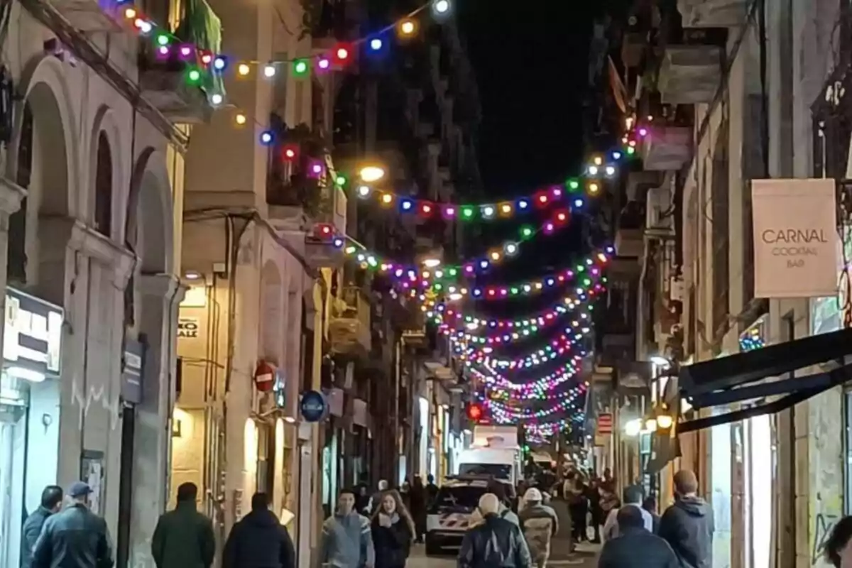 Una calle iluminada por luces de colores con personas caminando por la acera y edificios a ambos lados.