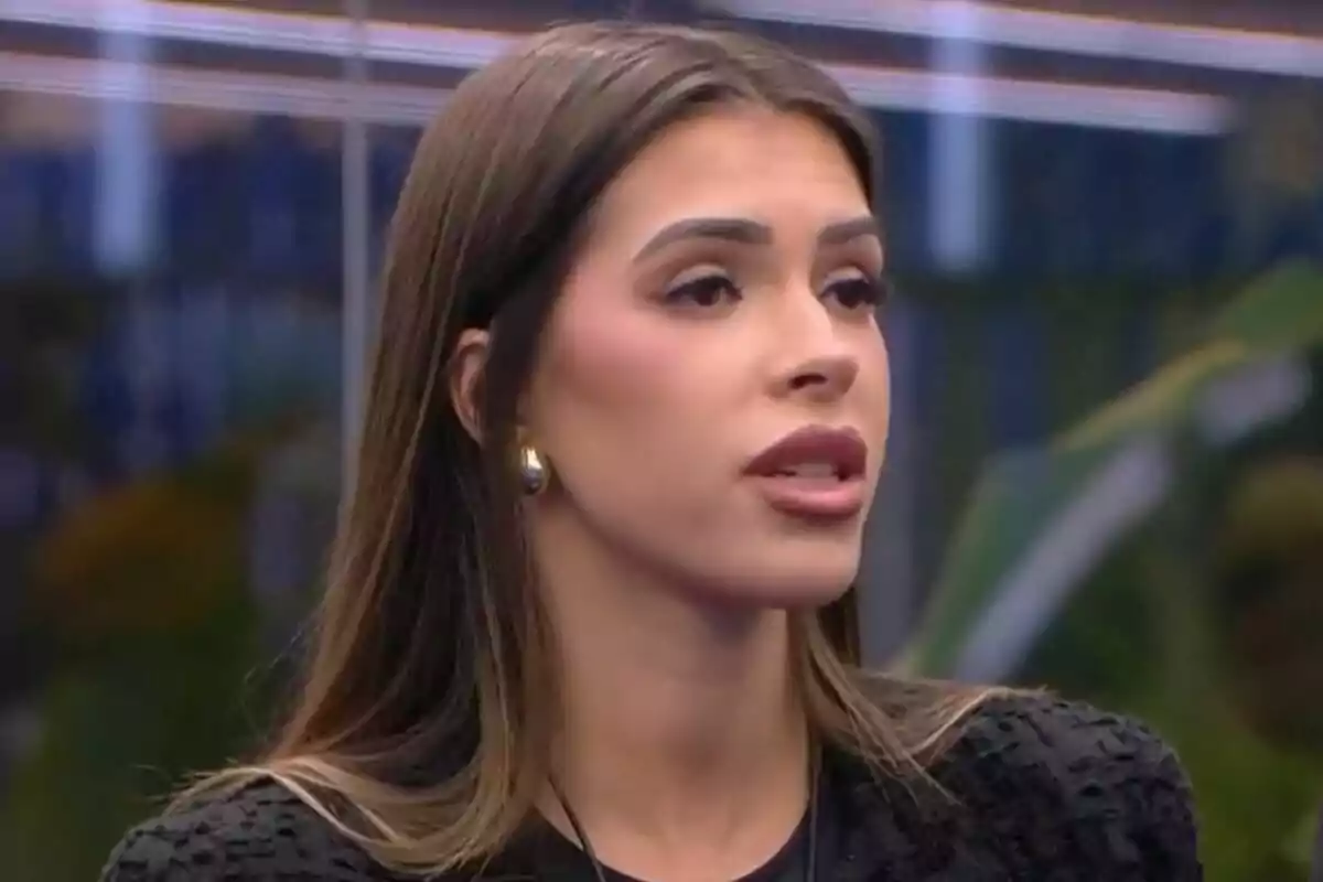 Lucía Rolek cabello largo y oscuro con expresión seria, usando aretes dorados y un atuendo negro, en un entorno interior en Gran Hermano.