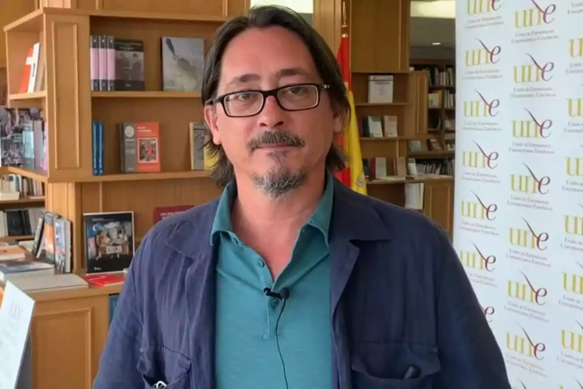 Luis Felipe Torrente con gafas y barba está de pie en una biblioteca, con estanterías llenas de libros detrás de él y un cartel de la Unión de Editoriales Universitarias Españolas (UNE) a su derecha.