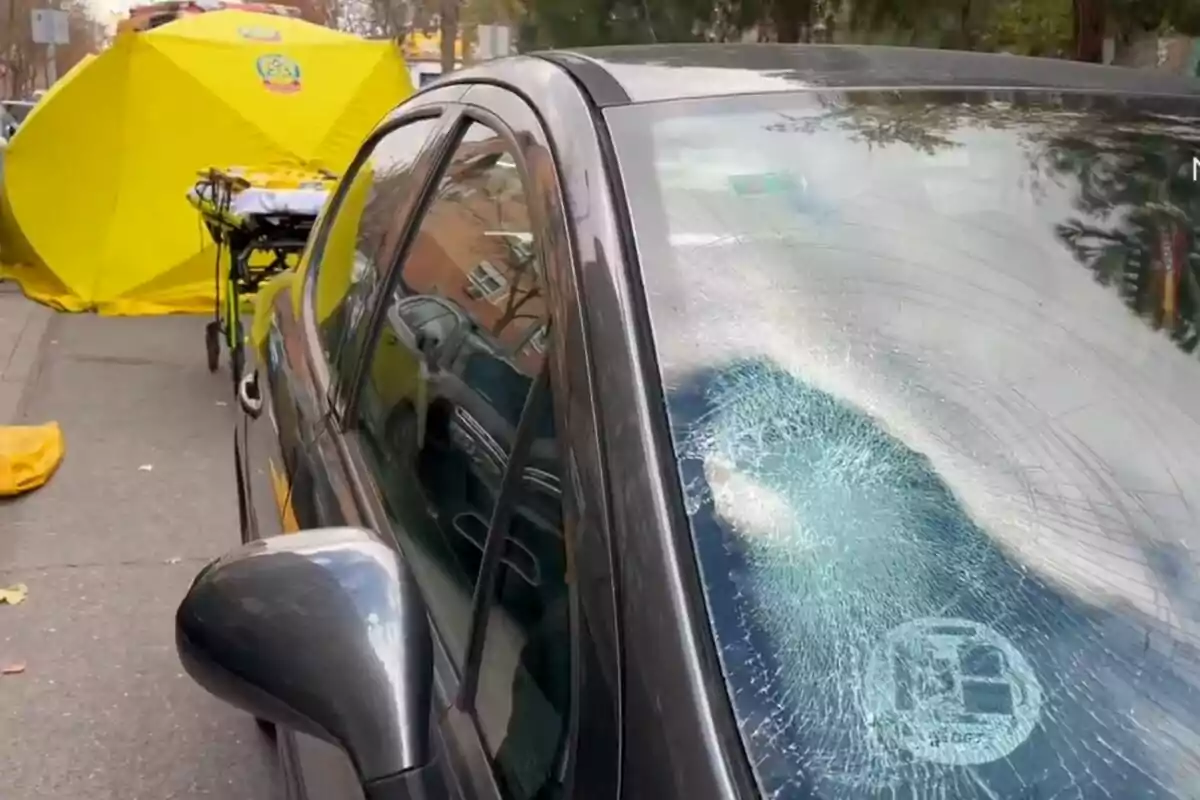Un coche negro con el parabrisas roto está estacionado en una calle junto a una carpa amarilla y una camilla. Un coche negro con el parabrisas roto está estacionado en una calle junto a una carpa amarilla y una camilla.