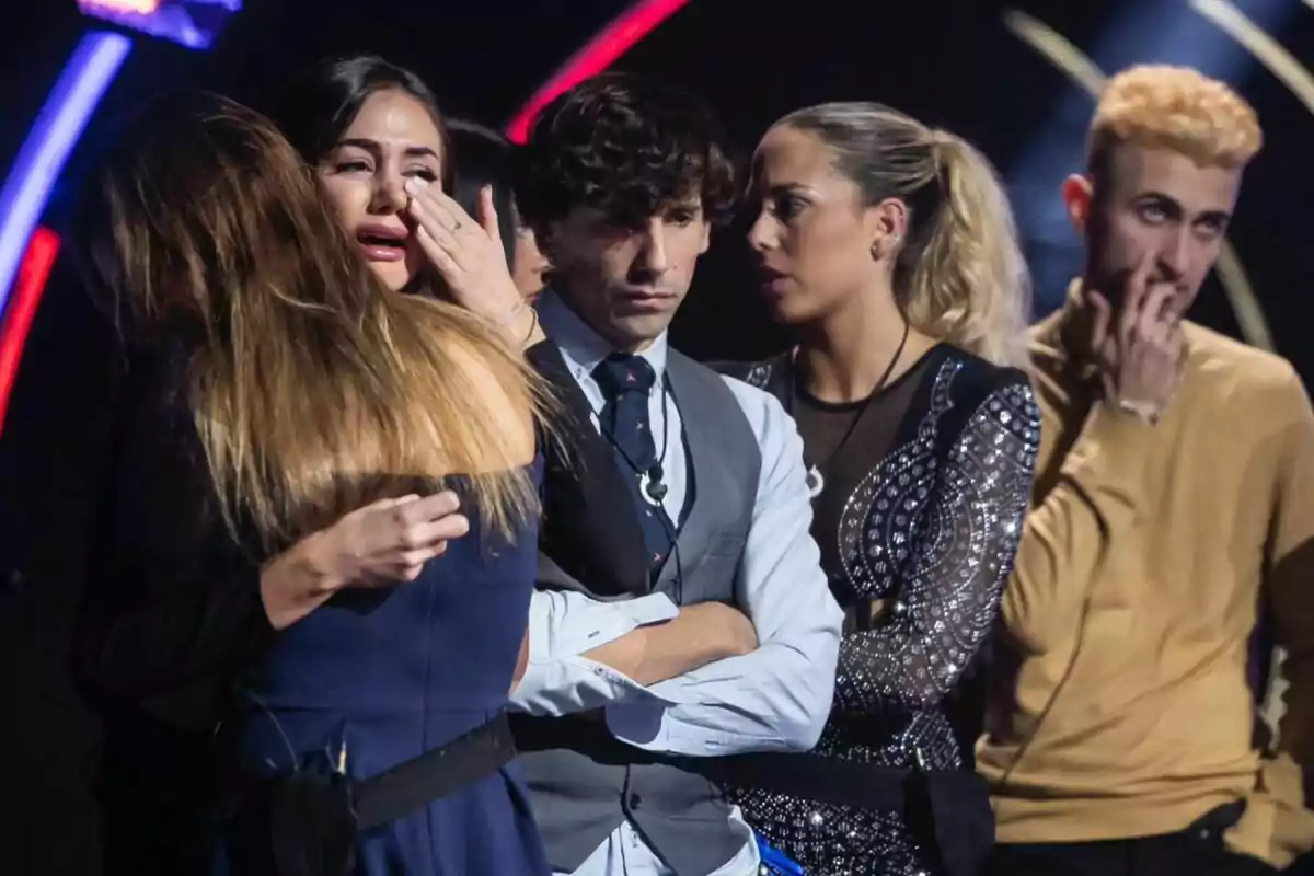 Un grupo de personas en un escenario, Maica está llorando mientras es abrazada por otra persona, y los demás observan con expresiones serias en la casa de Gran Hermano.