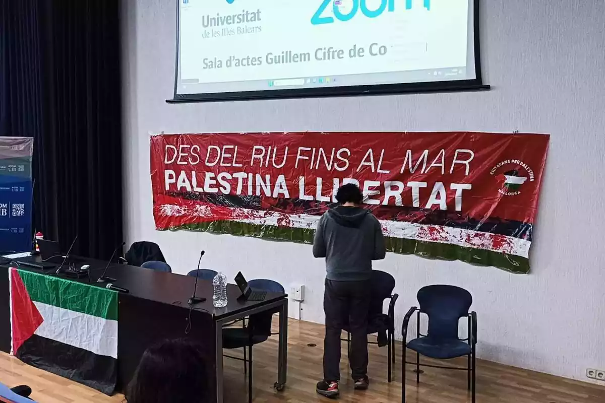 Sala de la Universitat de les Illes Balears con una bandera palestina en la mesa de los conferenciantes y de fondo un pancarta con el lema "Palestina libertad, desde el río hasta el mar"