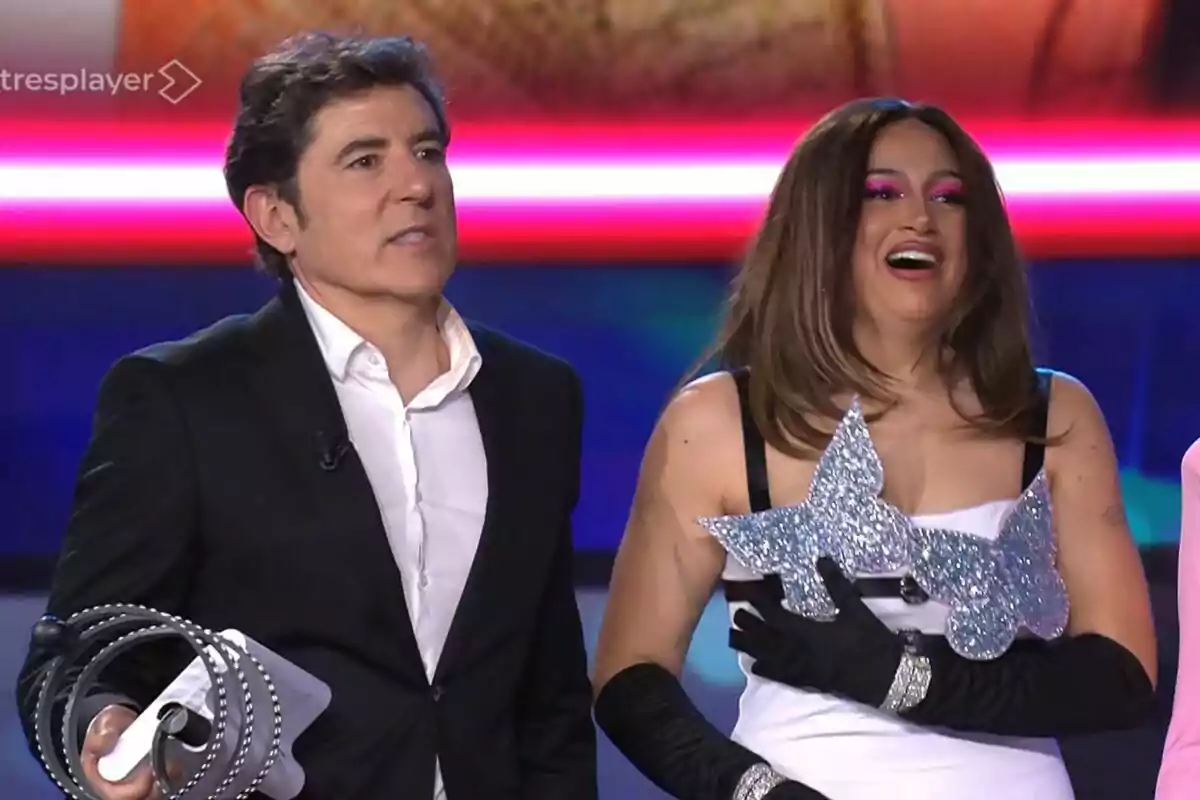 Manel Fuentes y Esperansa Grasia en un escenario, una de ellas sostiene un objeto brillante en forma de mariposa mientras sonríe, y la otra lleva un traje formal oscuro en Tu Cara Me Suena. Manel Fuentes y Esperansa Grasia en un escenario, una de ellas sostiene un objeto brillante en forma de mariposa mientras sonríe, y la otra lleva un traje formal oscuro en Tu Cara Me Suena.
