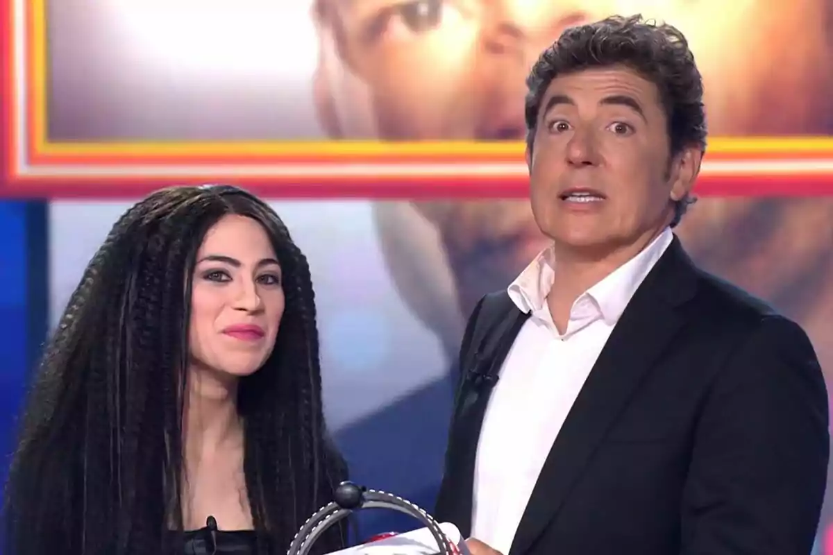 Melani García y Manel Fuentes en un set de televisión de Tu Cara Me Suena, una mujer con cabello largo y rizado y un hombre con traje oscuro y camisa blanca, ambos mirando hacia la cámara.