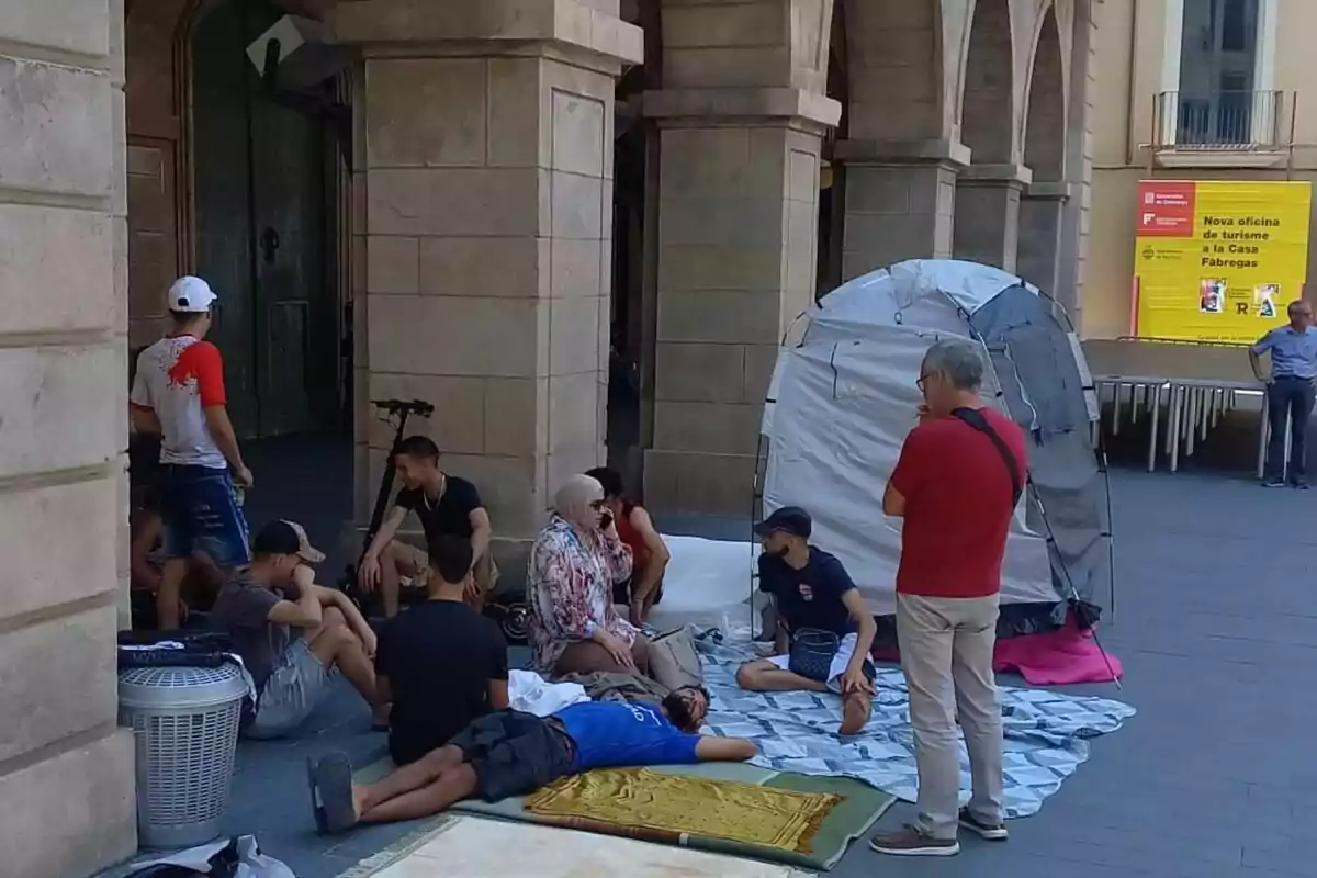 Un grupo de personas se encuentra reunido en una plaza, algunas sentadas y otras acostadas sobre mantas, junto a una tienda de campaña y un edificio de piedra con arcos.