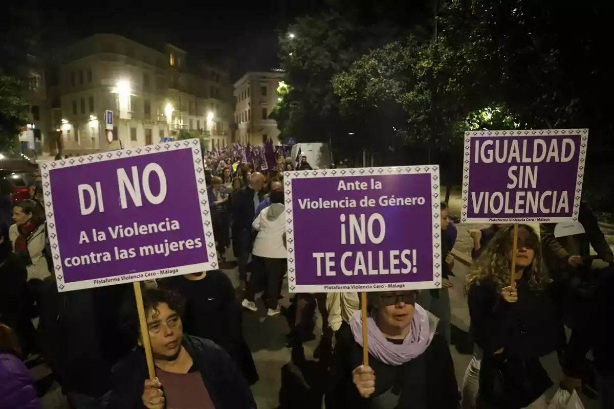 Personas marchando de noche con pancartas moradas que dicen "Di no a la violencia contra las mujeres", "Ante la violencia de género no te calles" e "Igualdad sin violencia".