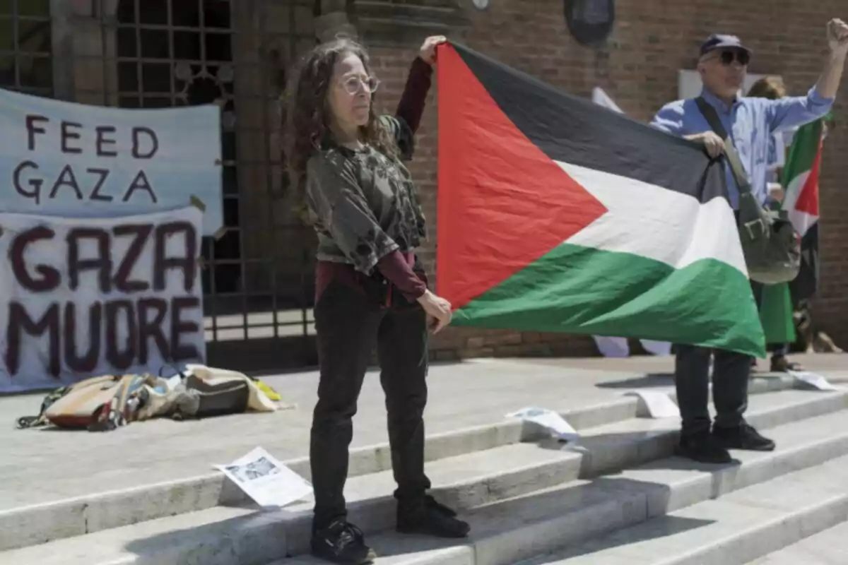 Mujer sosteniendo una bandera de Palestina durante una manifestación, con pancartas de apoyo a Gaza al fondo Mujer sosteniendo una bandera de Palestina durante una manifestación, con pancartas de apoyo a Gaza al fondo