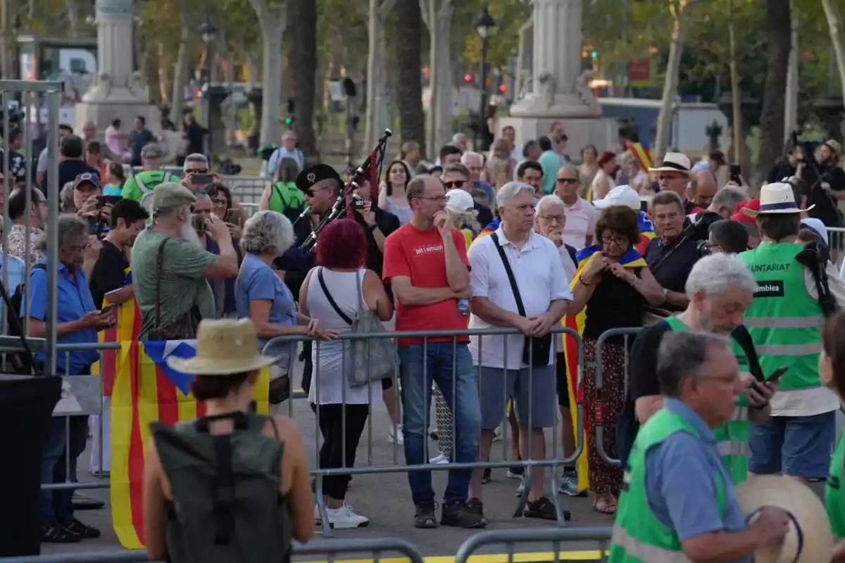 Un grupo de personas se encuentra reunido en un espacio público, algunas de ellas portando banderas y otras tomando fotos, mientras un hombre toca la gaita y varios voluntarios con chalecos verdes están presentes.