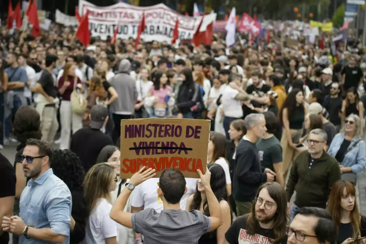 Una multitud de personas participa en una manifestación, algunas portan pancartas y banderas, mientras un individuo sostiene un cartel que dice "Ministerio de Supervivencia".