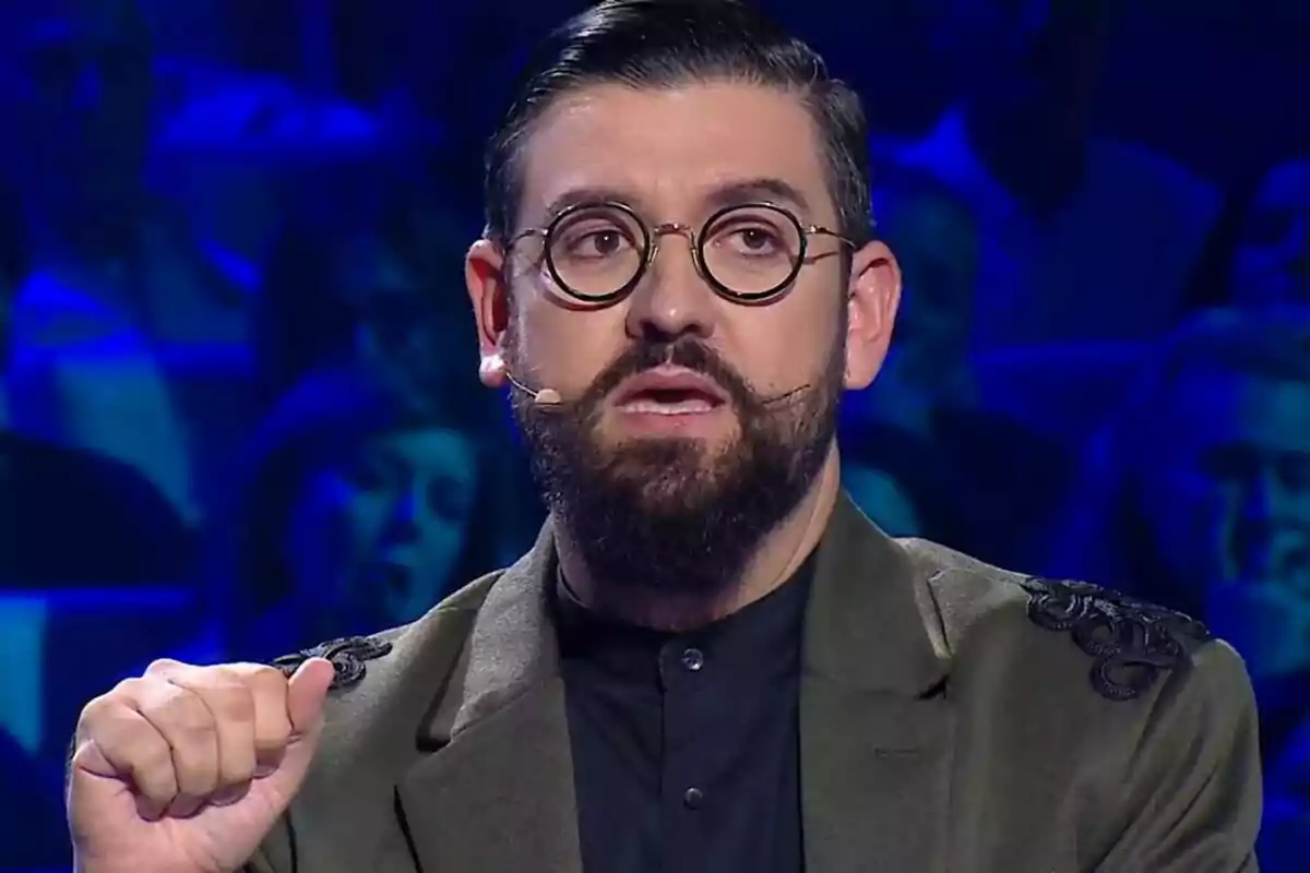 Manu Sánchez con gafas redondas y barba hablando en un escenario iluminado en azul en Somos Música.